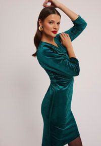 Turkusowa aksamitna sukienka z długimi rękawami, dekoltem w serek i marszczeniami po bokach, połączona z dużymi złotymi kolczykami.