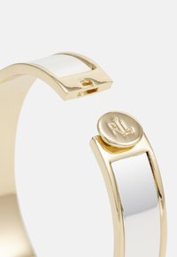 Gullfarget mansjettarmbånd med en glatt hvit emaljeinnlegg, rektangulær form og en rund logo på den ene enden.