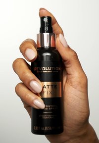 Bouteille de spray noire avec un accent métallique rose doré. L'étiquette du produit indique "Matte Fix" et "Makeup Revolution". Main tenant la bouteille.
