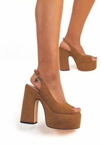 Dei tacchi a piattaforma in suede marrone con design open-toe, una fascia sul retro e un tacco a blocco spesso con un dettaglio lucido.