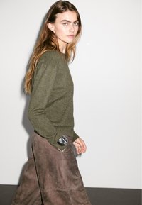 Maglione a maglia verde oliva con scollo a giro, abbinato a pantaloni larghi in suede marrone con dettagli sulle tasche.