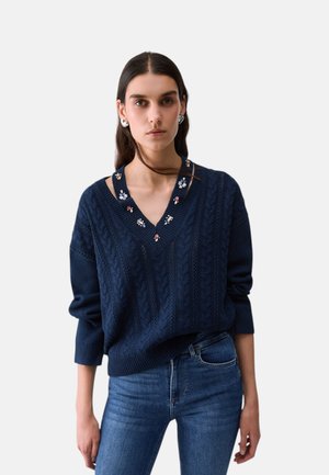 Giovane donna con lunghi capelli scuri indossa un maglione blu navy a trecce con ricamo floreale e jeans blu, in piedi davanti a uno sfondo bianco.