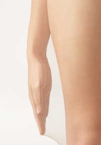 Průsvitné nahé punčochy s jemnou síťovinou, hladce obepínající nohu. Ruka je jemně položena na oblasti stehna, což zvýrazňuje přiléhavost.