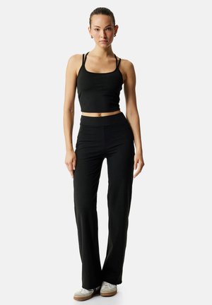 Nuori nainen seisoo mustassa lyhyessä topissa, korkeavyötäröisissä mustissa housuissa ja valkoisissa tennareissa yksiväristä taustaa vasten.