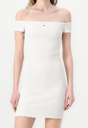 Femme portant une mini-robe blanche texturée à épaules dénudées avec un petit logo sur la poitrine, debout devant un fond uni.