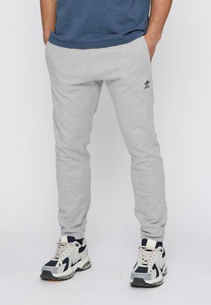 Graue Baumwoll-Sweatpants mit einem figurbetonten Design, ausgestattet mit einem gestickten schwarzen Logo, kombiniert mit weißen und dunkelblauen Sportschuhen.