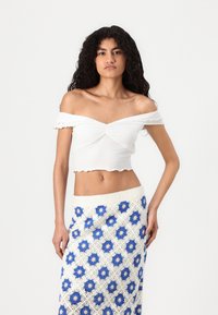 Hvit av-shoulder crop top med volanger og tvinnet detalj, parret med et mønstret blått og hvitt heklet skjørt.
