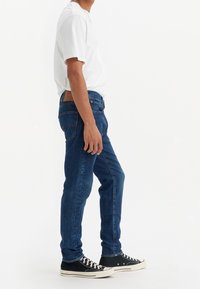 Siniset denim-farkut slim fit -mallilla, viidellä taskulla varustettuna ja takana ruskea nahkapatch. Yhdistetty valkoiseen t-paitaan ja mustiin lenkkikenyihin.