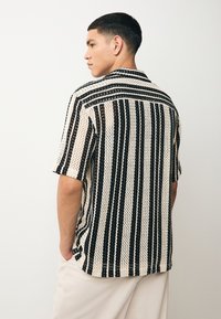 Camisa de manga corta hecha de tela de punto abierto en negro y crema, con rayas verticales y un corte holgado, con cuello puntiagudo y dobladillo suelto.