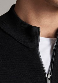 Pull en tricot noir avec fermeture éclair, col montant, tissu texturé et détail de fermeture éclair argentée à l'avant.