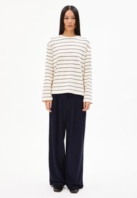 Langärmliges gestreiftes Oberteil in Creme mit schwarzen Linien, kombiniert mit lockeren, navyfarbenen Hosen mit weitem Bein und weicher Textur. Schwarze Schuhe.