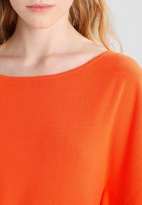 Jaskrawo pomarańczowa bluzka z długim rękawem, o szerokim wycięciu w kształcie łódki i miękkiej, teksturowanej tkaninie. Zbliżenie ukazuje szczegóły koloru i materiału.