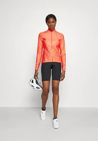 Γυναίκα που φοράει φωτεινό πορτοκαλί ποδηλατικό μπουφάν με φερμουάρ, μαύρο ποδηλατικό σορτς, άσπρα ποδηλατικά παπούτσια και κρατάει ένα λευκό κράνος στο πλάι της.