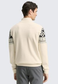 Cremfarbener Pullover mit hohem Kragen und gemusterten schwarzen Ärmeln mit geometrischen Designs. Glatte Stricktextur, taillierte Passform.