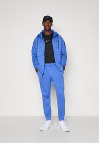 Blue Nike tracksuit with zip-up hoodie and joggers, black Nike t-shirt underneath, and white sneakers; fitted, smooth textures, subtle logo accents.