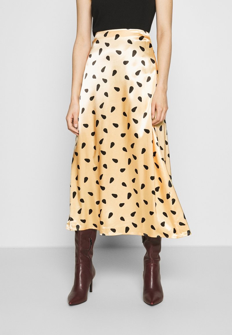 Jupe midi en satin couleur or pâle avec un motif de gouttes noires, présentant un design en cache-cœur. Associée à des bottes bordeaux jusqu'aux genoux.