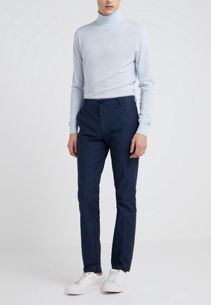 Mann trägt hellblauen Rollkragenpullover, marineblaue schmale Hose und weiße Sneakers, steht vor einem einfarbigen hellgrauen Hintergrund.