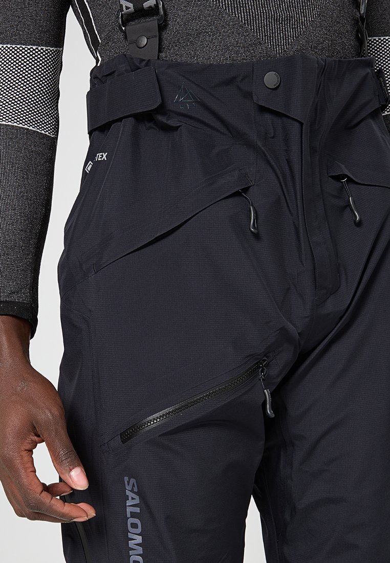Zwarte waterdichte salopette met verstelbare banden, ritszakken en gestructureerde stof. Beschikt over een logo op het been en een hoge taille.
