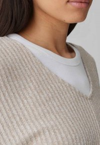 Beiger, gerippter Pullover mit V-Ausschnitt, der über einem weißen Rundhalsausschnitt-Shirt getragen wird. Strukturierter Stoff mit vertikalen Linien, der ein weiches Erscheinungsbild erzeugt.