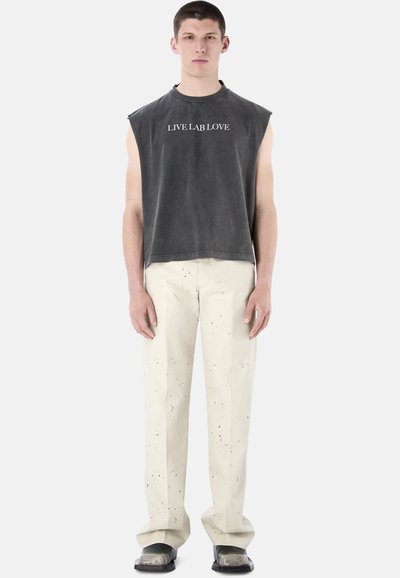 Joven con camiseta sin mangas negra desgastada con el texto "LIVE LAB LOVE", pantalones beige moteados y zapatos negros, posando contra un fondo blanco.
