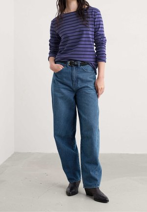 Woman wearing blue and black striped long-sleeve shirt, high-waisted loose blue jeans with black belt, and black ankle boots, standing indoors.