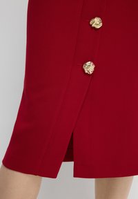 Rode kokerrok met een fitted ontwerp, voorzien van goudkleurige knopen en een zijsplit voor gemakkelijke beweging. Glad, textuurstof.