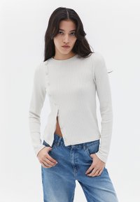 Top a maniche lunghe di un beige chiaro a costine con chiusure a bottone laterali, abbinato a jeans blu. Il materiale sembra morbido con una vestibilità aderente.