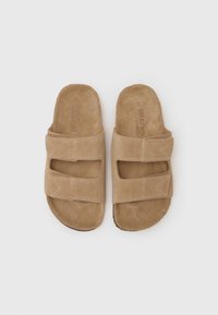 Beige suede papucs két állítható pánttal, lekerekített orral és lapos talppal, diszkrét varrással és puha textúrával.