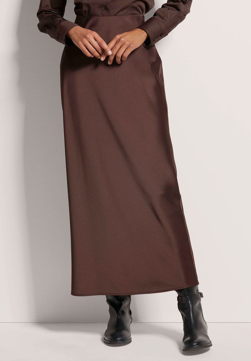 Bruine, vloerlange rok van gladde stof met een strakke structuur. Draagt bij zwarte, nauwsluitende enkellaarsjes en een knoopshirt.