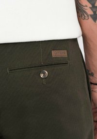 Olivgröna byxor med en texturerad tyg, som har en bakficka, en knappstängning och en läderlapp präglad med "ombre."