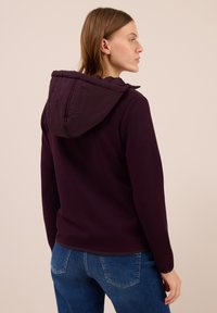 Femme de profil à droite portant une veste à capuche violet foncé et un jean bleu sur un fond beige uni.