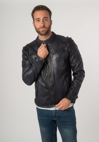 Zwarte leren jas met een staande kraag, ritszakken en textuurdetails op de schouders. Draagbaar over een grijs overhemd en blauwe jeans.