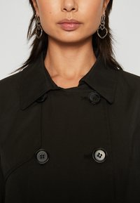 Cappotto nero con design doppiopetto, grandi bottoni e colletto classico. Abbinato a orecchini a forma di cuore.