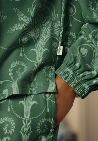Zaļš ziedu raksta audums ar sarežģītiem dizainiem un brīvu piegriezumu, elastīgām aprocēm un mazu etiķeti sānos.