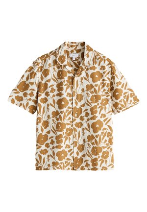 Mand stående i beige bukser, hvide sneakers og en kortærmet skjorte med knapper med et brunt blomsterprint på en lys baggrund.