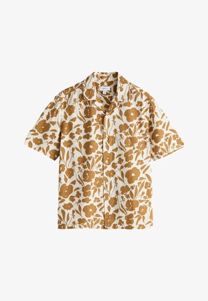 Mand stående i beige bukser, hvide sneakers og en kortærmet skjorte med knapper med et brunt blomsterprint på en lys baggrund.