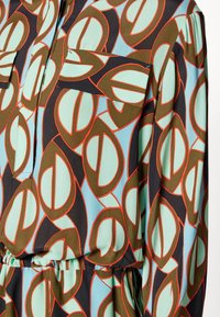 Chemise à manches longues avec motif géométrique en vert, bleu clair, marron et orange, avec poche poitrine et détail taille froncée.