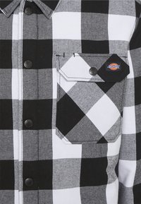 Chemise à carreaux noir et blanc avec une poche poitrine boutonnée portant une étiquette de la marque Dickies.