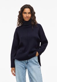 Marinefarbener Rollkragenpullover mit lockerer Passform, gerippten Bündchen und seitlichen Schlitzen. Kombiniert mit hellblauen Denim-Jeans. Weicher Stricktextur.