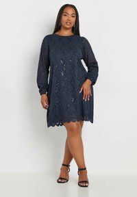 Vestido de encaje azul marino con mangas largas, dobladillo festoneado y sutiles detalles de lentejuelas, combinado con unos tacones negros con tiras.