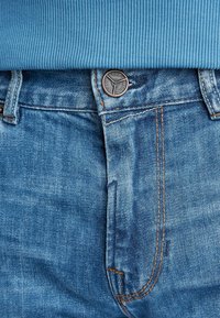 Modra jeans jakna iz jeansa z vidnim šivanjem in bronastim gumbom v pasu. Tkanina ima svetlo pranje in rahlo obrabljen videz.