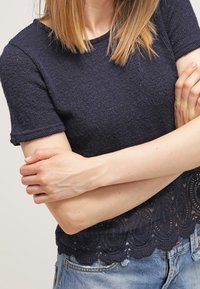 Námořnický top s krátkým rukávem, s texturovaným pleteným designem a vlnitým krajkovým lemem. Má volný střih a vrstvený látkový detail ve spodní části.