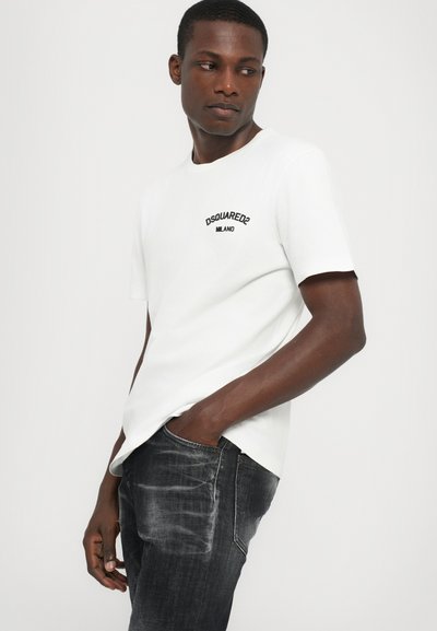 Camiseta blanca de algodón de manga corta, con un logotipo bordado en negro en el lado izquierdo del pecho. Combinada con unos vaqueros oscuros desgastados.
