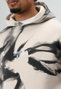 Sweatshirt met een beige basis, zwarte abstracte patronen, een capuchon met trekkoord en een wit "PRESENTED" logo op de borst.