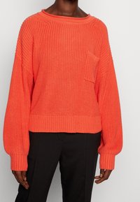 Korálový pletený svetr s volným střihem, širokým lodičkovým výstřihem, žebrovanými lemy na rukávech a přední kapsou. Vyrobený z texturované příze, kombinovaný s černými kalhotami.