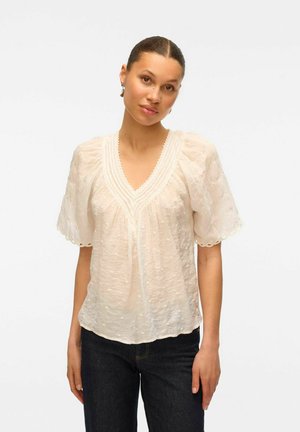 White, short-sleeve blouse in textured fabric, featuring a deep V-neck and decorative stitching. Worn over dark denim jeans.