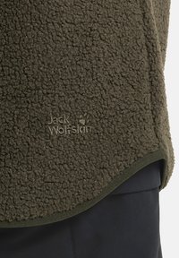 Fleecejacke in dunkelolivgrün mit einer strukturierten, plüschigen Oberfläche. Hat einen abgerundeten Saum und ein auf dem Stoff geprägtes Logo-Tag.