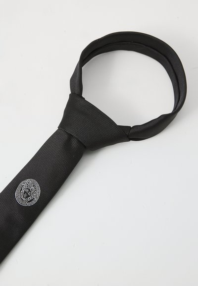 Cravate en soie noire avec une texture côtelée et un emblème argenté circulaire présentant un visage stylisé. La cravate est soigneusement nouée en haut.
