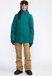 Vihreänsininen vedenpitävä takki hupulla, yhdistetty vaaleanruskeisiin lumihousuihin. Mustat käsineet ja saappaat. Lumilasit päässä.