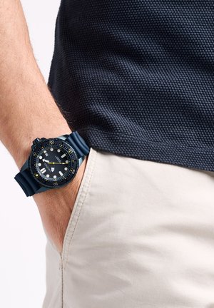 Zwarte duikhorloge met een gestructureerde bezel en witte uurmarkers. Blauwe silicium band. Gedig in de zak van een lichtgekleurde broek.